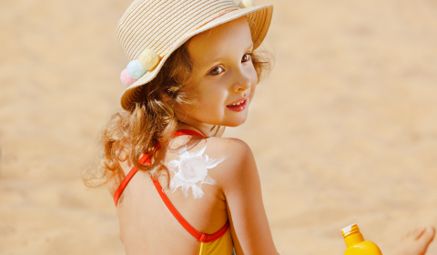 Lapsi hiekkarannalla hellehattu p&auml;&auml;ss&auml;, &auml;iti levitt&auml;&auml; aurinkovoidetta lapselle